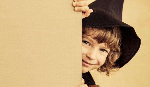 A girl peeking her head past a wall with a witch hat on